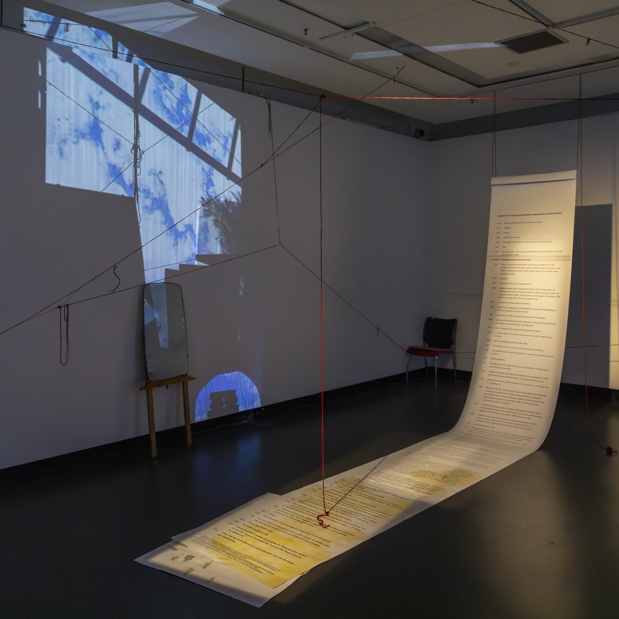 A long paper scroll of printed text rising from the floor to the ceiling beside projected images