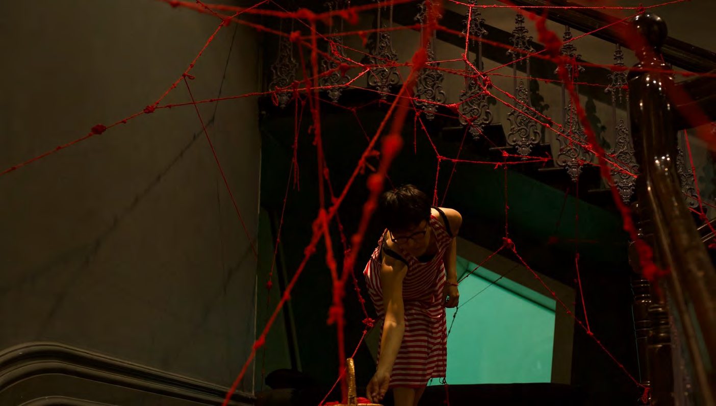 The artist weaving among red yarn in a dark gallery